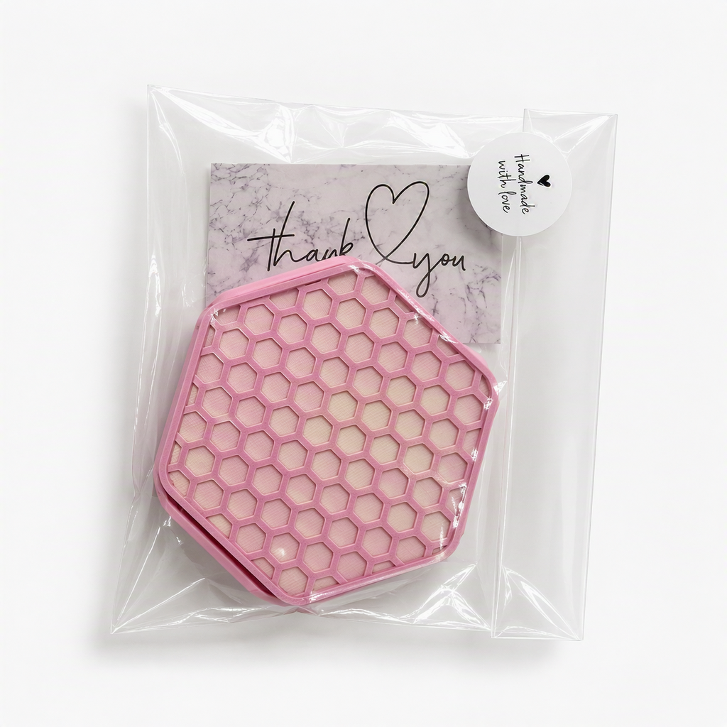 A pink hexagonal honeycomb-patterned Coaster set, with holder, sealed inside a clear plastic bag with a round white sticker that reads 'Handmade with love' and a small branded card behind it.